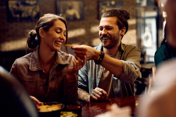 Young happy man feeding his girlfriend while eating in pub. - obrazy, fototapety, plakaty