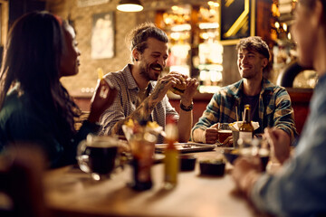 Cheerful man eating burger while gathering with friends in bar.