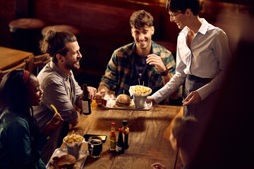Group of happy friends drinking beer while waitress is serving them food in pub.