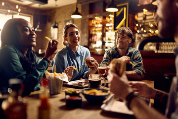 Multiracial group of happy friends eating burgers while drinking beer in pub. - obrazy, fototapety, plakaty