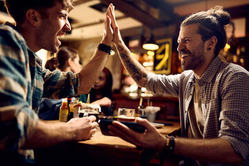 Cheerful men giving high-five while watching sports match on smart phone in a pub.