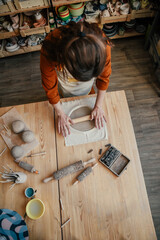 Artisan woman in lighted pottery studio, rolling out clay for modelling. Pottery studio with a variety of ceramics and a focused artisan , top view