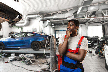 Pensive young African mechanic in car service looking at car thinking of how to repair it