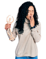 Young hispanic woman with curly hair holding small wooden manikin bored yawning tired covering mouth with hand. restless and sleepiness.