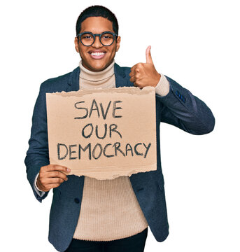 Young handsome hispanic man holding save our democracy protest banner smiling happy and positive, thumb up doing excellent and approval sign