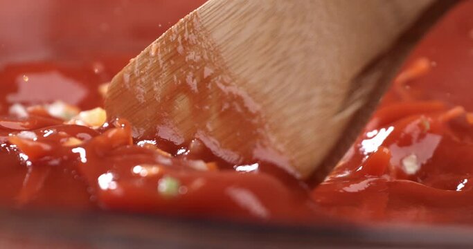Super slow motion macro of fresh organic bio tomato sauce with sautéed vegetables is being mixed with wooden spoon while cooking in pan for pasta dish preparation in restaurant kitchen.