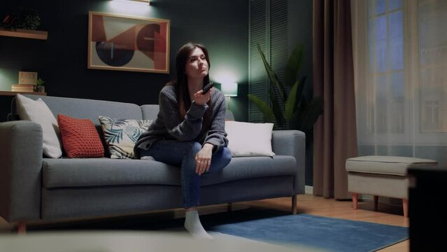 Woman sitting at couch in comfortable apartment. Sad female sitting at couch. Woman holding remote control. Female changing channels on TV set. Bored woman have nothing to watch.