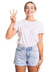 Young beautiful blonde woman wearing casual white tshirt showing and pointing up with fingers number three while smiling confident and happy.