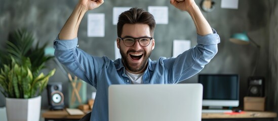 Young businessman rejoicing with raised arms at his desk, gazing at laptop screen.