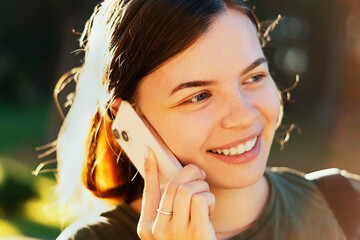 Happy, smiling young girl talking on a mobile phone in a city park. Young beautiful lady talking on her smartphone outdoors. An attractive girl happily communicates with a friend. Communication