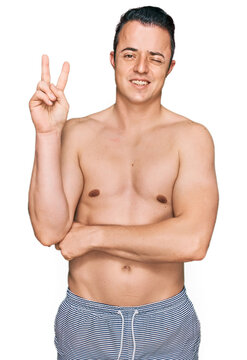 Handsome young man wearing swimwear shirtless smiling with happy face winking at the camera doing victory sign with fingers. number two.