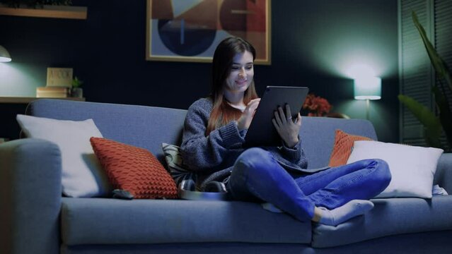 Smiling woman relaxing sitting at sofa. Happy female using gadget. Woman holding tablet and working. Female touching device screen. Cheerful woman looking at tablet. Staying at home.
