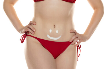 Young woman in red bikini with cosmetic product in smile shape on her belly on white background.