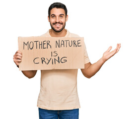 Young hispanic man holding mother nature is crying protest cardboard banner celebrating victory with happy smile and winner expression with raised hands