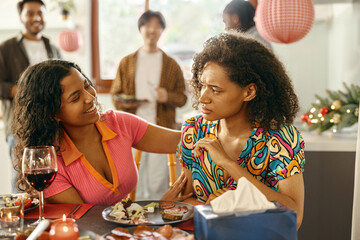 Positive female friend supporting upset young woman while sitting at home during party time