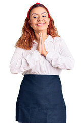 Young latin woman wearing waitress apron praying with hands together asking for forgiveness smiling confident.