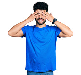 Young arab man with beard wearing casual blue t shirt covering eyes with hands smiling cheerful and funny. blind concept.