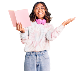 Young beautiful mixed race woman reading a book wearing headphones celebrating victory with happy smile and winner expression with raised hands