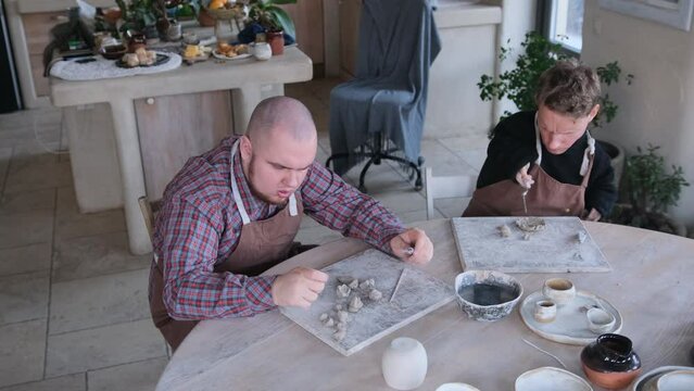 two young people with autism syndrome and with special physiological development are engaged in clay modeling sitting at a large table in a home workshop.