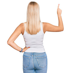 Young blonde girl wearing casual style with sleeveless shirt posing backwards pointing ahead with finger hand