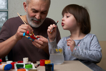 Grandfather and granddaughter paint a toy made of clay. Family leisure time.