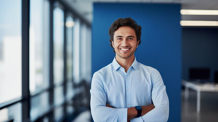 Obraz premium Cheerful man in a business casual blue shirt, arms confidently folded, stands in a contemporary office space, image suitable for themes of innovation, teamwork, and approachable leadership