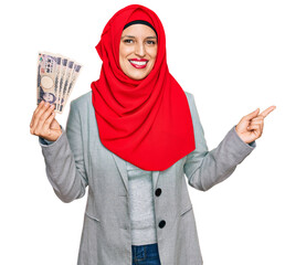 Beautiful hispanic woman wearing islamic hijab holding japanese yen banknotes smiling happy pointing with hand and finger to the side