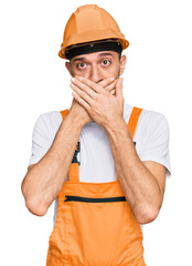 Hispanic young man wearing handyman uniform and safety hardhat shocked covering mouth with hands for mistake. secret concept.