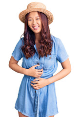 Young beautiful chinese girl wearing summer hat smiling and laughing hard out loud because funny crazy joke with hands on body.