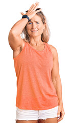 Middle age fit blonde woman wearing casual summer clothes and sunglasses surprised with hand on...