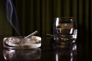 ashtray with a lit cigarette next to an empty glass on a table, smoking day, mysterious and melancholic atmospher