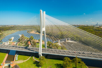 Long pylon bridge spanning Oder river against sunny Wroclaw. Cars drive on cable-stayed Redzinski Bridge among scenic city parks aerial view