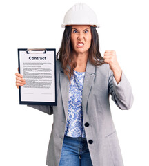 Young beautiful brunette woman wearing architect hardhat holding contract annoyed and frustrated shouting with anger, yelling crazy with anger and hand raised