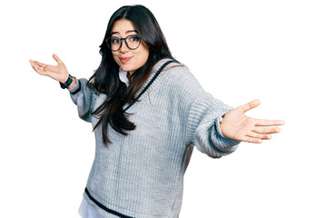 Beautiful hispanic woman wearing casual sweater and glasses clueless and confused expression with arms and hands raised. doubt concept.