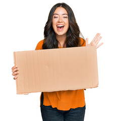 Beautiful brunette young woman holding cardboard blank empty banner celebrating victory with happy smile and winner expression with raised hands