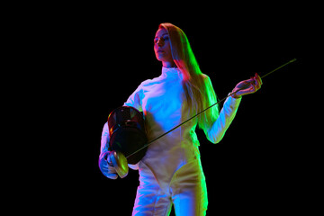 Portrait of young woman, fencer, athlete in white uniform posing with sword over black background...
