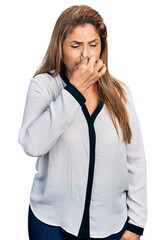 Middle age latin woman wearing business shirt smelling something stinky and disgusting, intolerable smell, holding breath with fingers on nose. bad smell