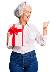 Senior grey-haired woman holding gift pointing thumb up to the side smiling happy with open mouth