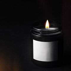 A lit decorative paraffin candle in a black glass jar on a black background. There is a blank white...