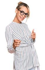 Beautiful blonde woman wearing business shirt and glasses pointing fingers to camera with happy and funny face. good energy and vibes.