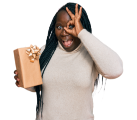 Young black woman with braids holding gift smiling happy doing ok sign with hand on eye looking through fingers