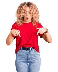 Young blonde woman with curly hair wearing casual red tshirt pointing down with fingers showing advertisement, surprised face and open mouth - obrazy, fototapety, plakaty