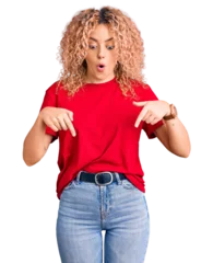 Foto op Canvas Young blonde woman with curly hair wearing casual red tshirt pointing down with fingers showing advertisement, surprised face and open mouth © Krakenimages.com