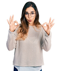 Beautiful hispanic woman wearing casual sweater and glasses looking surprised and shocked doing ok approval symbol with fingers. crazy expression