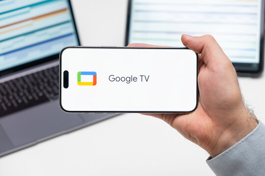 Google TV Logo Of App On The Screen Of Mobile Phone Held By Man In Front Of The Laptop And Tablet, December 2023, Prague, Czech Republic