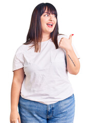 Young plus size woman wearing casual clothes smiling with happy face looking and pointing to the side with thumb up.