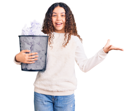 Beautiful kid girl with curly hair holding paper bin full of crumpled papers celebrating victory with happy smile and winner expression with raised hands