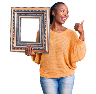 Young african american woman holding empty frame pointing thumb up to the side smiling happy with open mouth