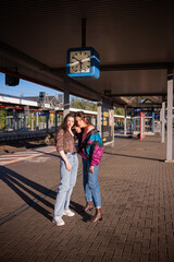 Two beautiful girls dressed in 90s style posing on the platform of the train station under a clock...