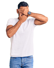Fototapeta premium Young latin man wearing casual clothes covering eyes and mouth with hands, surprised and shocked. hiding emotion